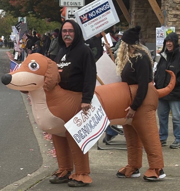 Two people riding blow up dog with signs saying No Kings and Save Democracy 